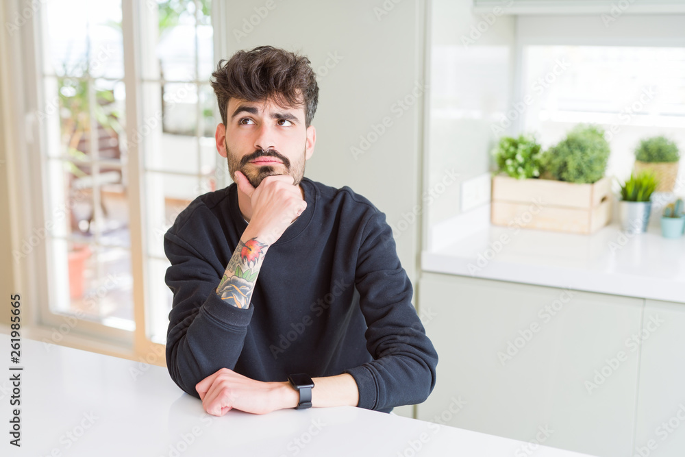 Young man wearing casual sweatshirt sitting on white table with hand on chin thinking about question, pensive expression. Smiling with thoughtful face. Doubt concept. Young man wearing casual sweatshirt sitting on white table with hand on chin thinking about question, pensive expression. Smiling with thoughtful face. Doubt concept.