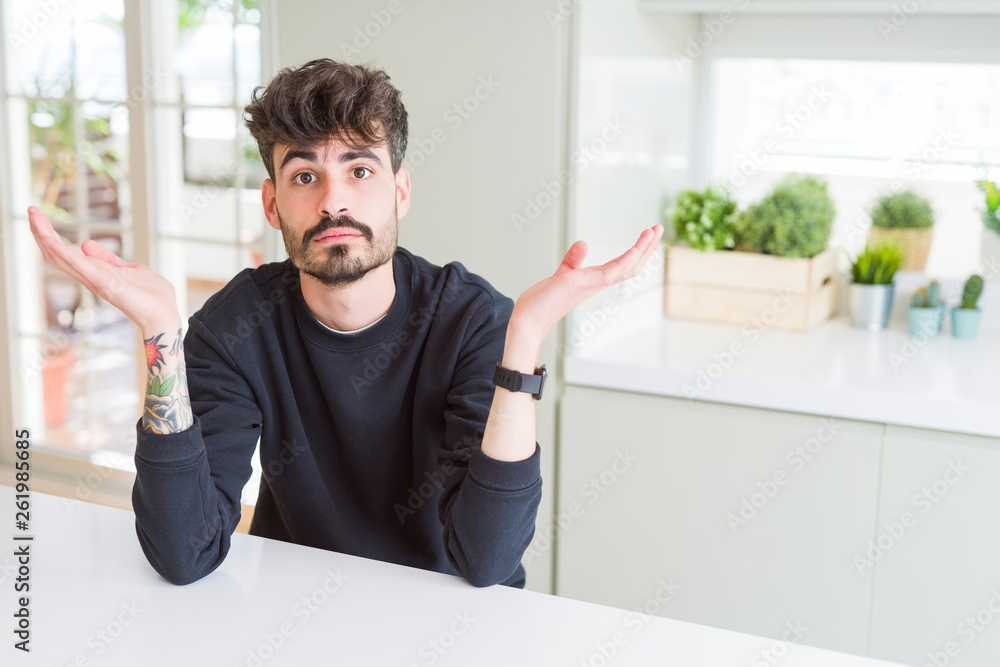 Young man wearing casual sweatshirt sitting on white table clueless and confused expression with arms and hands raised. Doubt concept.