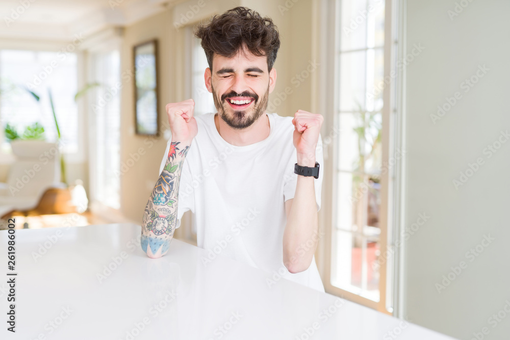 Young man wearing casual t-shirt sitting on white table excited for success with arms raised celebrating victory smiling. Winner concept. Young man wearing casual t-shirt sitting on white table excited for success with arms raised celebrating victory smiling. Winner concept.