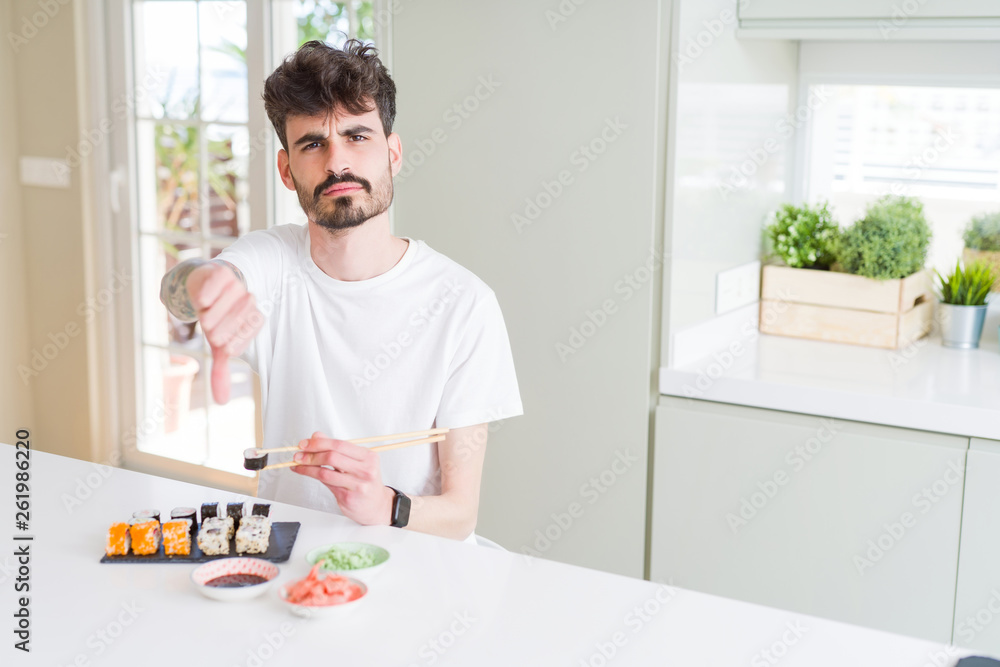 Young man eating asian sushi from home delivery with angry face, negative sign showing dislike with thumbs down, rejection concept Young man eating asian sushi from home delivery with angry face, negative sign showing dislike with thumbs down, rejection concept
