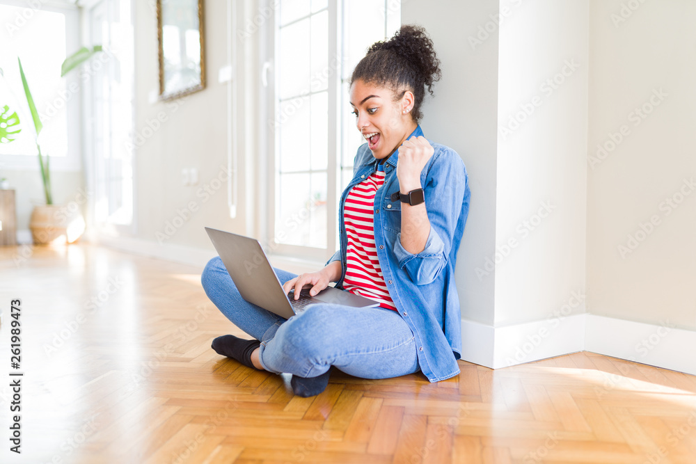 Young african american woman sitting on the floor using computer laptop screaming proud and celebrating victory and success very excited, cheering emotion