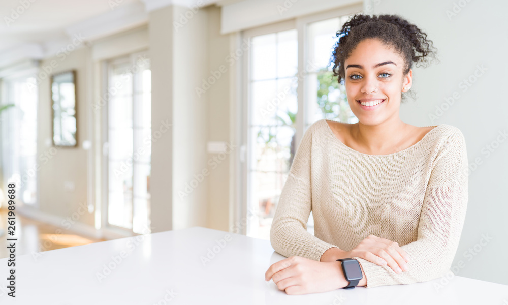 Beautiful young african american woman with afro hair sitting on table at home with a happy and cool smile on face. Lucky person. Beautiful young african american woman with afro hair sitting on table at home with a happy and cool smile on face. Lucky person.