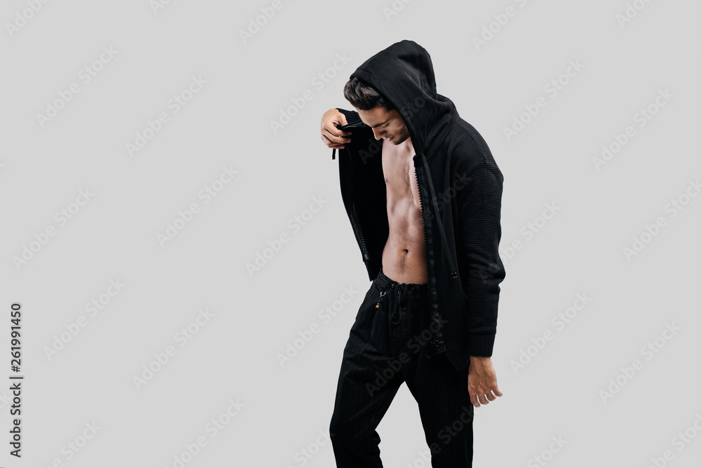 Stylish young dancer dressed in a sweatshirt on a naked torso with a hood and black pants stands on a white background Stylish young dancer dressed in a sweatshirt on a naked torso with a hood and black pants stands on a white background