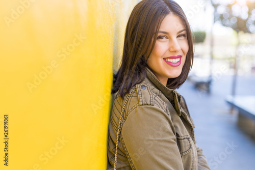 Beautiful young woman smiling confident and cheerful leaning on yellow wall, walking on the street of the city on a sunny day