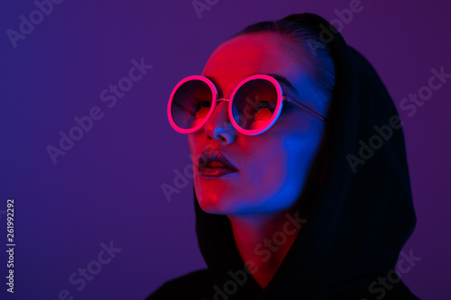 Portrait of fashion young girl in a black sweater with a hood and round sunglasses in red and blue neon light in the studio photo