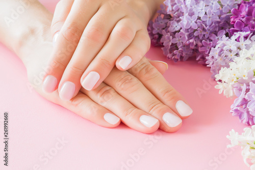 Young, perfect woman's hands with light nails on pastel pink table. Care about clean, soft, smooth skin. Manicure, pedicure beauty salon. Beautiful branches of lilac blossoms. Colorful, fresh flowers.