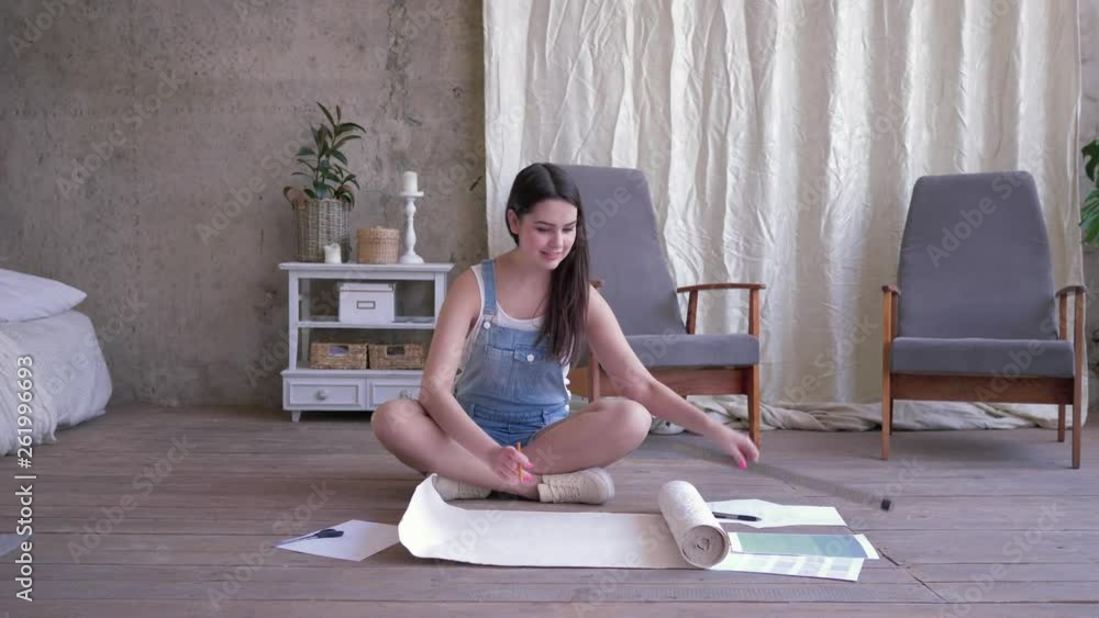 young girl unrolling a piece of white wallpaper makes pencil marks and then cuts off a piece with scissors sitting on floor in a new room. young girl unrolling a piece of white wallpaper makes pencil marks and then cuts off a piece with scissors sitting on floor in a new room.