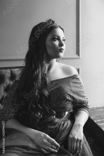monochrome (black and white) portrait of beautiful young pretty cute woman princess (queen) in long purple queen's dress and crown, with long hair and make up indoor in minimalistic interior