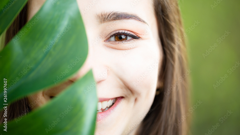 Beautiful natural young woman closeup behind big monstera leaf with green background in the woods. Beautiful natural young woman closeup behind big monstera leaf with green background in the woods.
