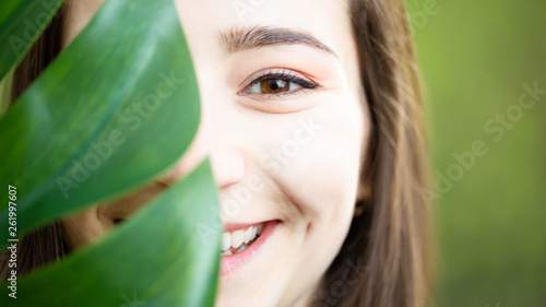 Beautiful natural young woman closeup behind big monstera leaf with green background in the woods.