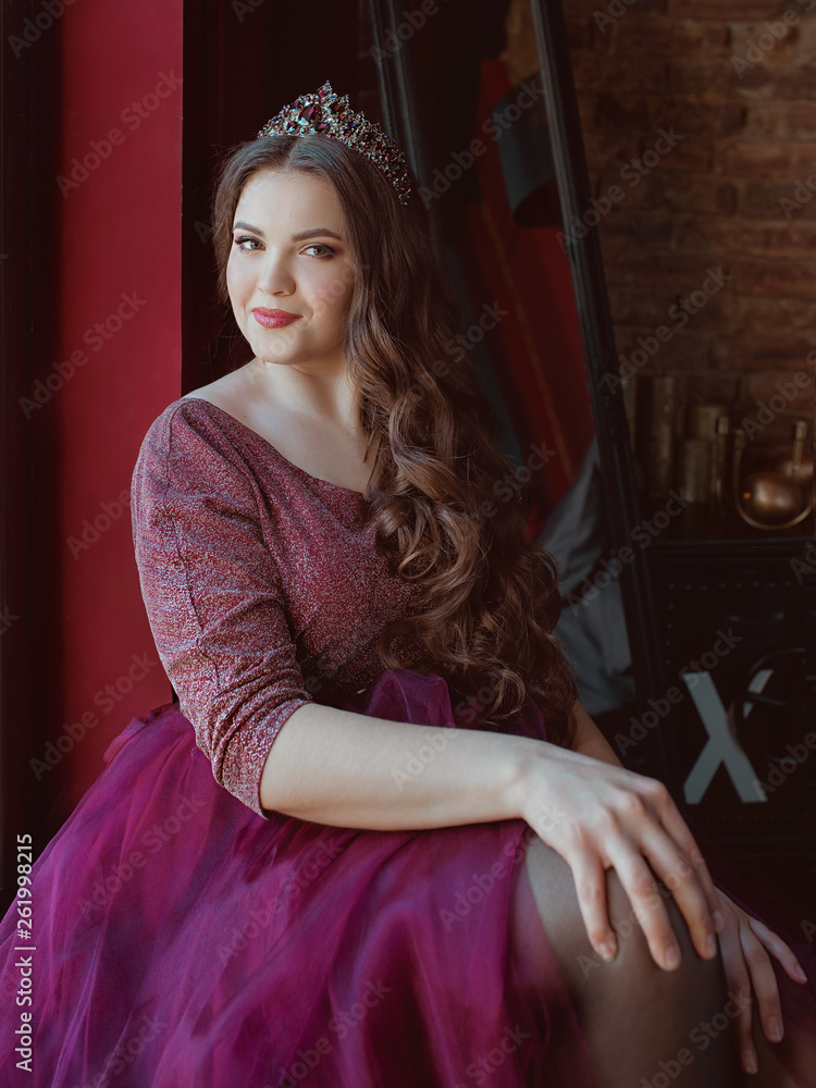 portrait of beautiful young pretty cute woman princess (queen) in long purple queen's dress and crown, with long hair and make up indoor in minimalistic loft interior sitting by the window