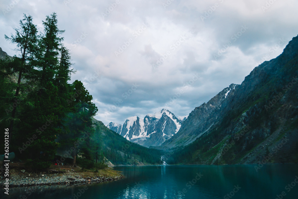 Wonderful mountain lake with view on giant glacier. Amazing huge mountains with conifer forest. Larch tree on water edge. Morning landscape of majestic nature of highlands. Cloudy mountainscape.