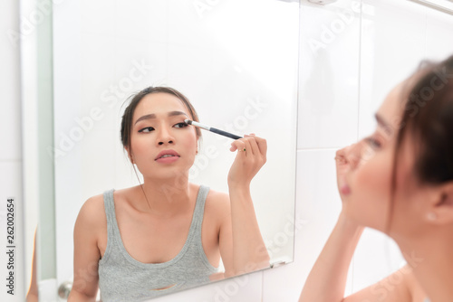 A young woman is standing in front of the bathroom mirror and putting on makeup.