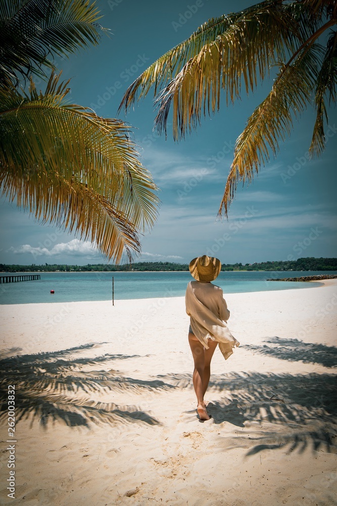 Woman in Tropical Look and Straw Hat walking on white sand tropical island beach. Summer Beach Look. Woman in Tropical Look and Straw Hat walking on white sand tropical island beach. Summer Beach Look.