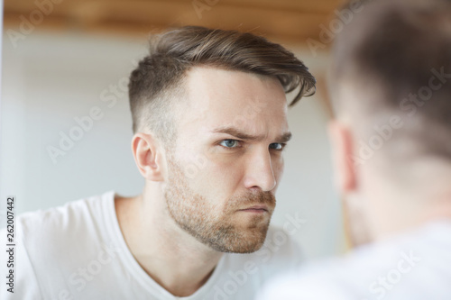 Head and shoulders portrait of handsome young  looking at his reflection in mirror and frowning, copy space