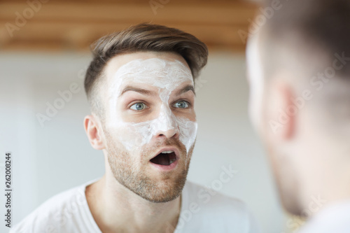 Head and shoulders portrait of handsome young man making faces applying cream and looking at his reflection in mirror, copy space