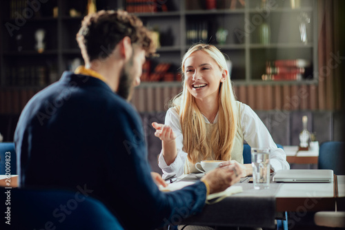 Smiling Caucasian blonde businesswoman dressed smart casual discussing with her male colleague about project while sitting in coffe shop.