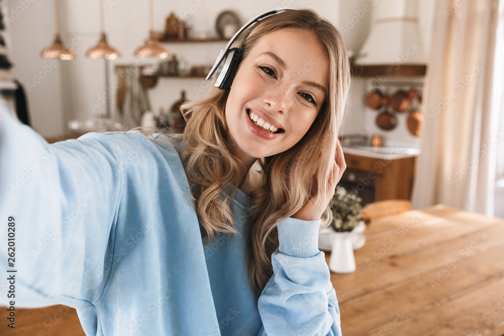 Portrait of pleased blond girl 20s wearing headphones smiling and taking selfie photo at home Portrait of pleased blond girl 20s wearing headphones smiling and taking selfie photo at home