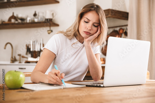 Image of disappointed blond woman studying on laptop and writing down notes while sitting at wooden table in apartment