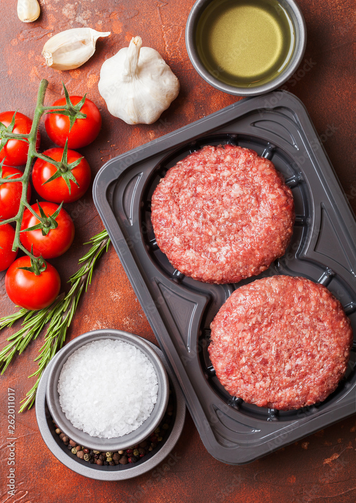 Plastic tray with raw minced homemade beef burgers with spices and herbs. Top view.On rusty kitchen table background with tomatoes salt and pepper. Plastic tray with raw minced homemade beef burgers with spices and herbs. Top view.On rusty kitchen table background with tomatoes salt and pepper.