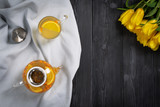 Transparent mug and teapot with sea buckthorn tea and yellow tulips on a dark wooden background.