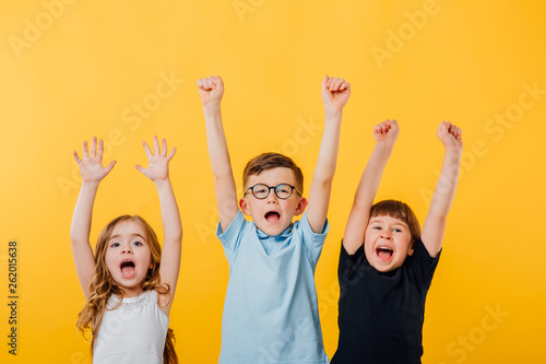 three children, a little boy and two little girls jumping up, their raised hands are happy, isolated yellow background, copy space photo
