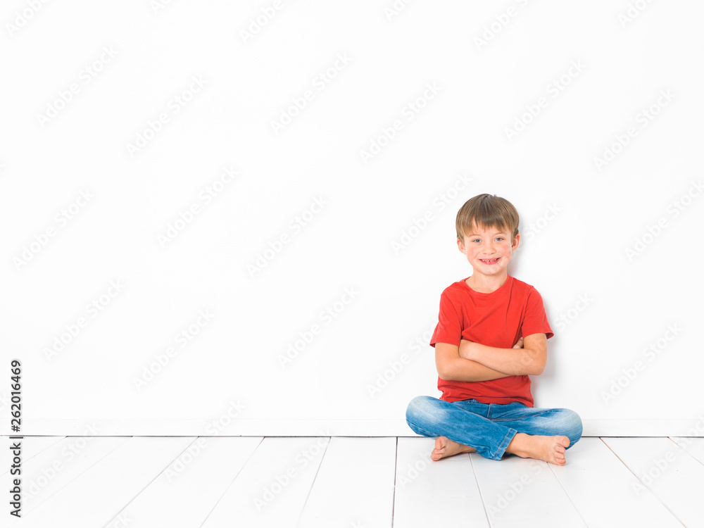 cute and blond boy with red shirt and blue jeans is posing on white wooden floor in front of white background cute and blond boy with red shirt and blue jeans is posing on white wooden floor in front of white background