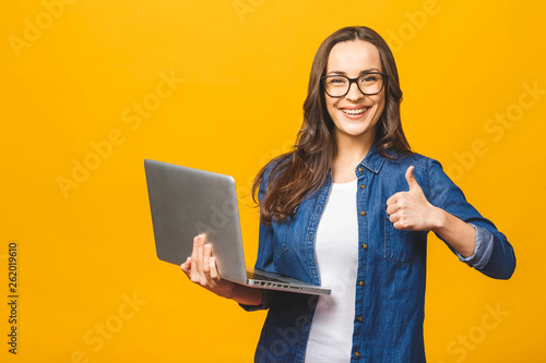 Portrait of a smiling young beautiful girl holding laptop computer and showing thumbs up isolated over yellow background.