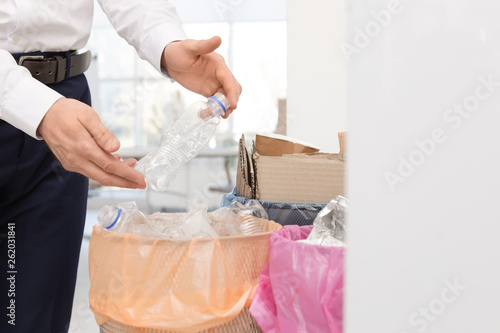 Man putting used plastic bottle into trash bin in office, closeup. Waste recycling