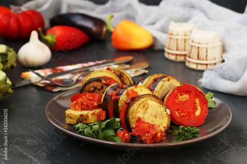 Ratatouille with baked vegetables: aubergines, zucchini and tomatoes is located on a plate on a dark background