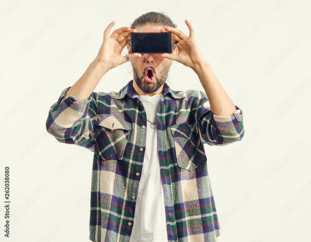 Portrait of a surprised male holding a smartphone in front of her eyes. Man closed her eyes with a mobile phone, standing on a light background. Social media, network, Internet - concept. Portrait of a surprised male holding a smartphone in front of her eyes. Man closed her eyes with a mobile phone, standing on a light background. Social media, network, Internet - concept.