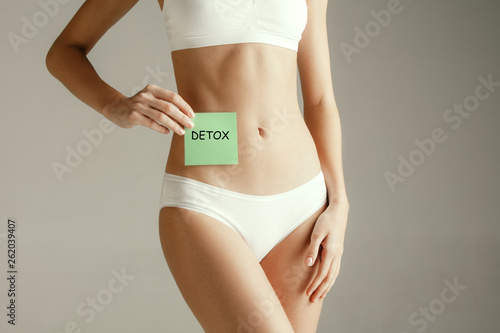 Young adult woman holding card with word DETOX. Woman health. Female model holding card near stomach isolated on gray studio background. Cut out part of body. Medical problem and solution. photo