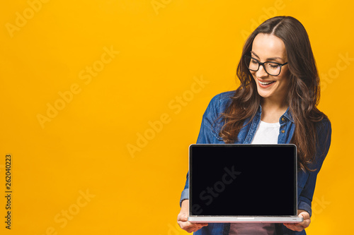 Surprised happy brunette woman in casual showing blank laptop computer screen and pointing on it while looking at the camera with open mouth over yellow background. photo