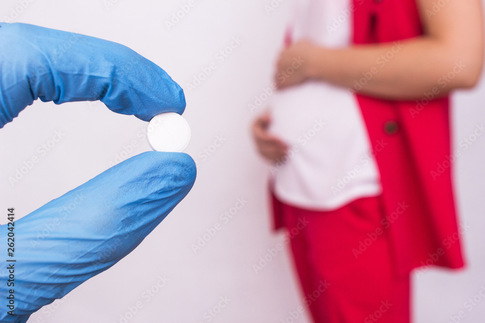 The doctor holds a pill in the form of a complex of vitamins and folic acid on the background of a pregnant girl, lack of vitamins and calcium during pregnancy The doctor holds a pill in the form of a complex of vitamins and folic acid on the background of a pregnant girl, lack of vitamins and calcium during pregnancy