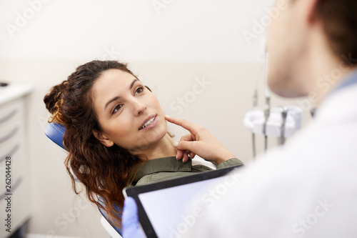 Portrait of beautiful young woman sitting in dentists chair and pointing at tooth while consulting with doctor, copy space