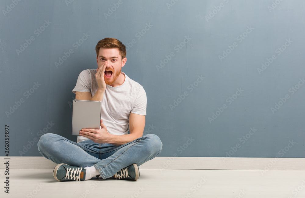 Young redhead student man sitting on the floor shouting something happy to the front. He is holding a tablet. Young redhead student man sitting on the floor shouting something happy to the front. He is holding a tablet.