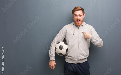 Young redhead fitness man surprised, feels successful and prosperous. He is holding a soccer ball.