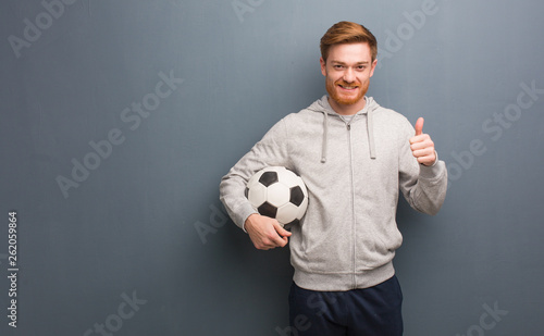 Young redhead fitness man smiling and raising thumb up. He is holding a soccer ball.