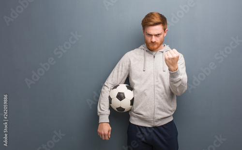 Young redhead fitness man showing fist to front, angry expression. He is holding a soccer ball.