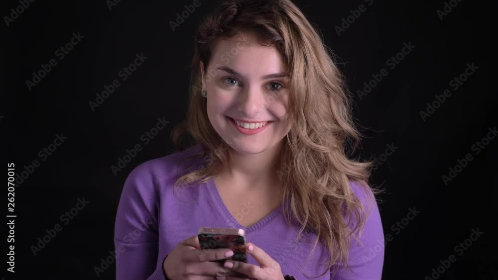 Portrait of glad woman working with smartphone watches into camera with kind smile on black background. Portrait of glad woman working with smartphone watches into camera with kind smile on black background.