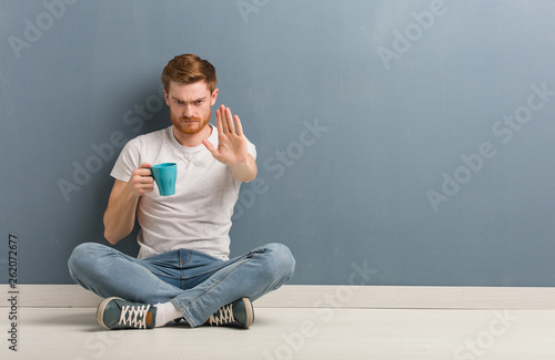 Young redhead student man sitting on the floor putting hand in front. He is holding a coffee mug.
