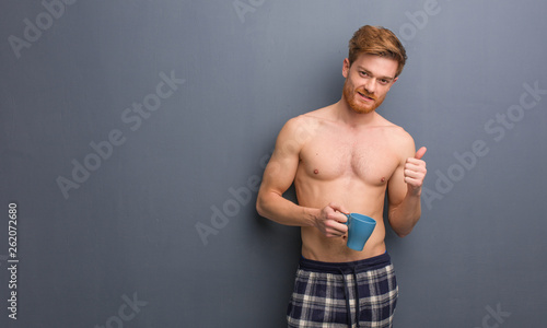Young shirtless redhead man smiling and raising thumb up. He is holding a coffee mug.