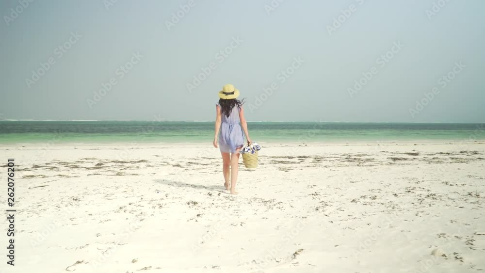 Young caucasian woman walking barefoot on sandy beach towards turquoise ocean coastline. Adult european girl in straw hat and sundress with basket in hand walks on deserted paradise beach at sunny day Young caucasian woman walking barefoot on sandy beach towards turquoise ocean coastline. Adult european girl in straw hat and sundress with basket in hand walks on deserted paradise beach at sunny day