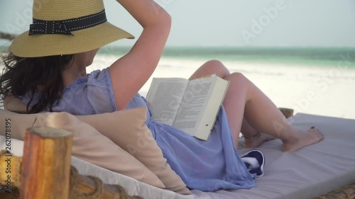 Young woman is reading book on white sand beach by ocean. Adult caucasian girl in straw hat and sundress is lying on lounger sunbed in shadow on white sandy beach and reading book at bright sunny day.