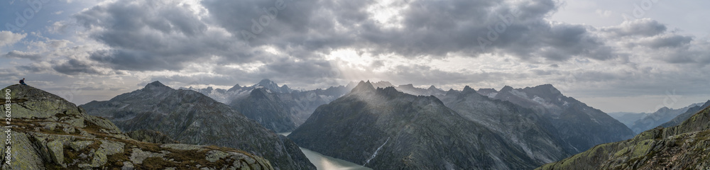 Panoramic view to the Grimsel lake in Swiss Alps with dramatic sun rays breaking through dark clouds with a boy resting on a rock