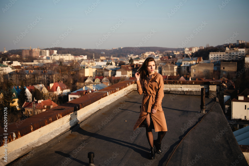 Young brunette woman in stylish dress posing on a rooftop with scenic view of the old city on sunset.