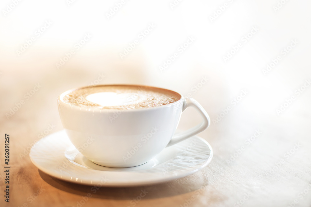 White cappuccino cup with with latte art heart on light brown wood background lit by bright morning sunlight with white copy space