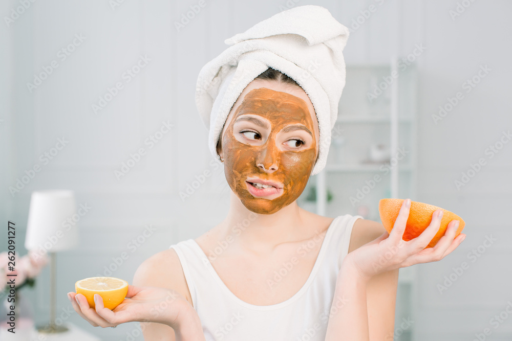 Funny beautiful woman holding lemon and grapefruit slices. Photo of girl in white towel with brown mud facial mask, having spa procedures. Funny beautiful woman holding lemon and grapefruit slices. Photo of girl in white towel with brown mud facial mask, having spa procedures.