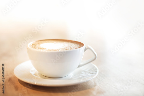 White cappuccino cup with with latte art heart on light brown wood background lit by bright morning sunlight with white copy space