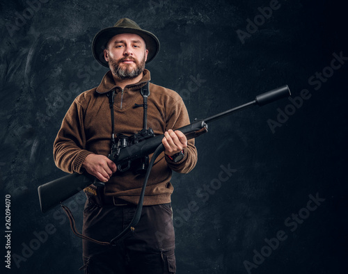 Mature hunter with rifle and binoculars. Studio photo against dark wall background